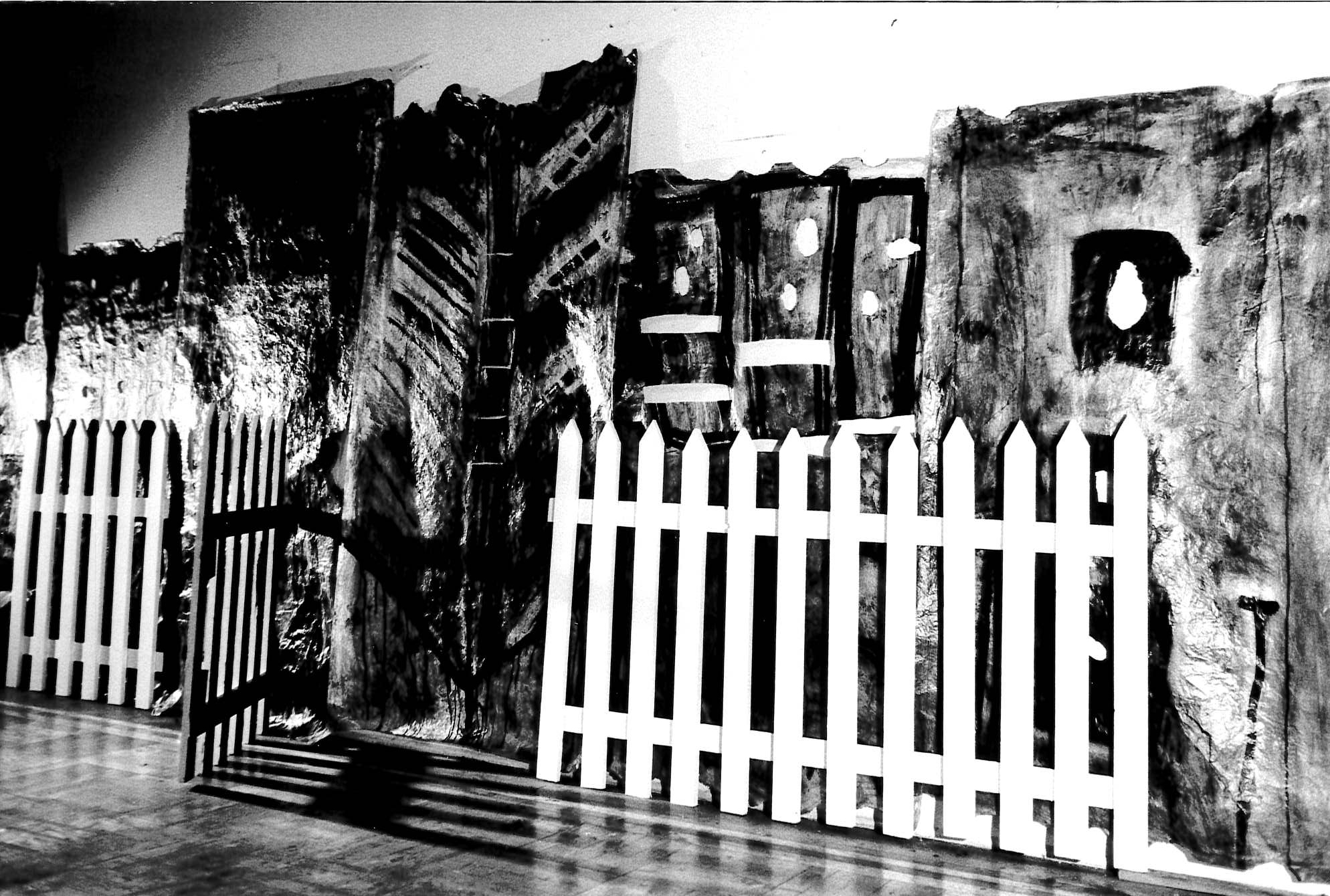 Black and white photograph of white picket fence opening with dark painted background behind it.