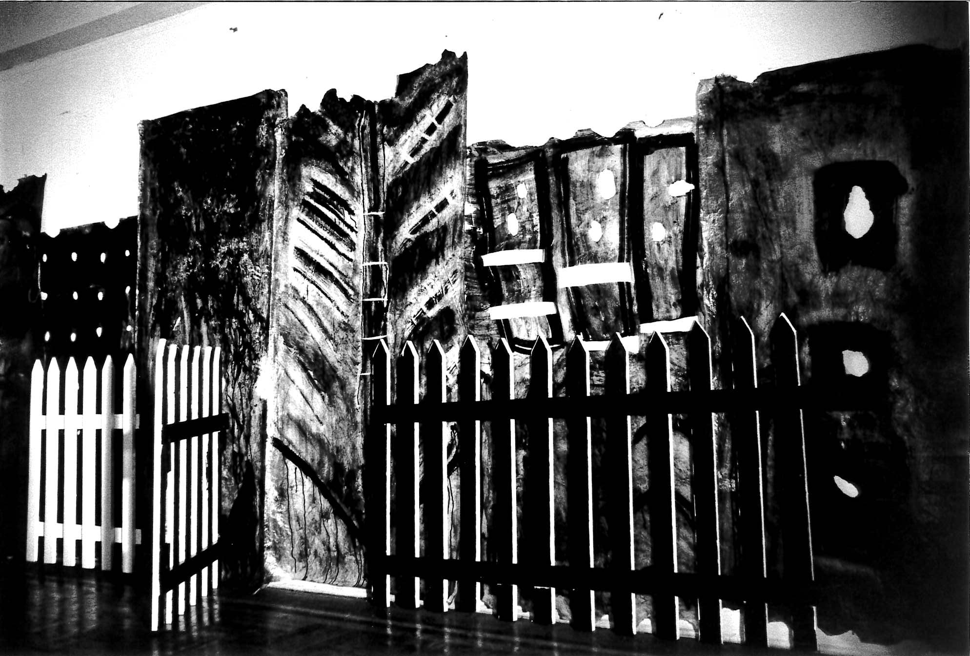 Black and white photograph of white picket fence with dark painted background behind it.