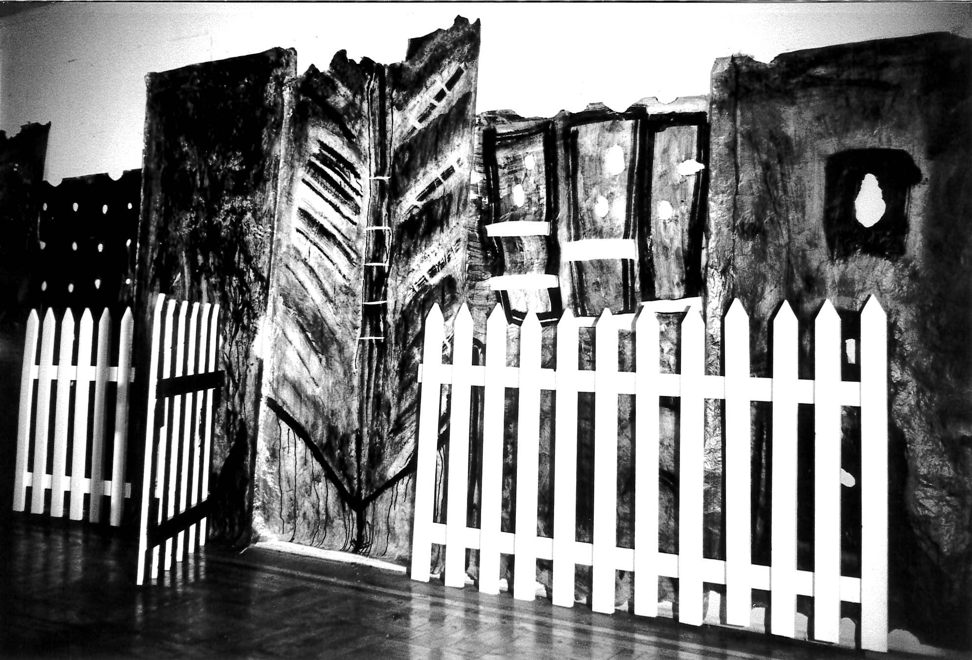Black and white photograph of white picket fence with dark painted background behind it.