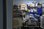 Color photograph of a room used to store medical props and devices, including hospital beds, catheters, and three medical test dummies stacked on top of one another on a shelf. The walls are painted a blue-grey color.
