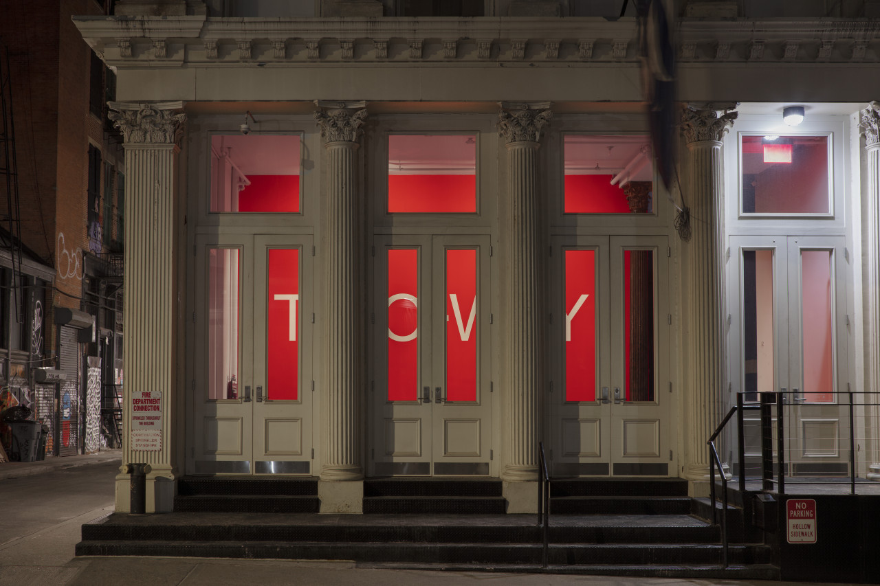 An exterior view of a gallery with white neoclassical columns and a row of three glass doors. Inside the gallery is a bright red wall with large white text: the text is partially obscured by the building’s columns and doorways.