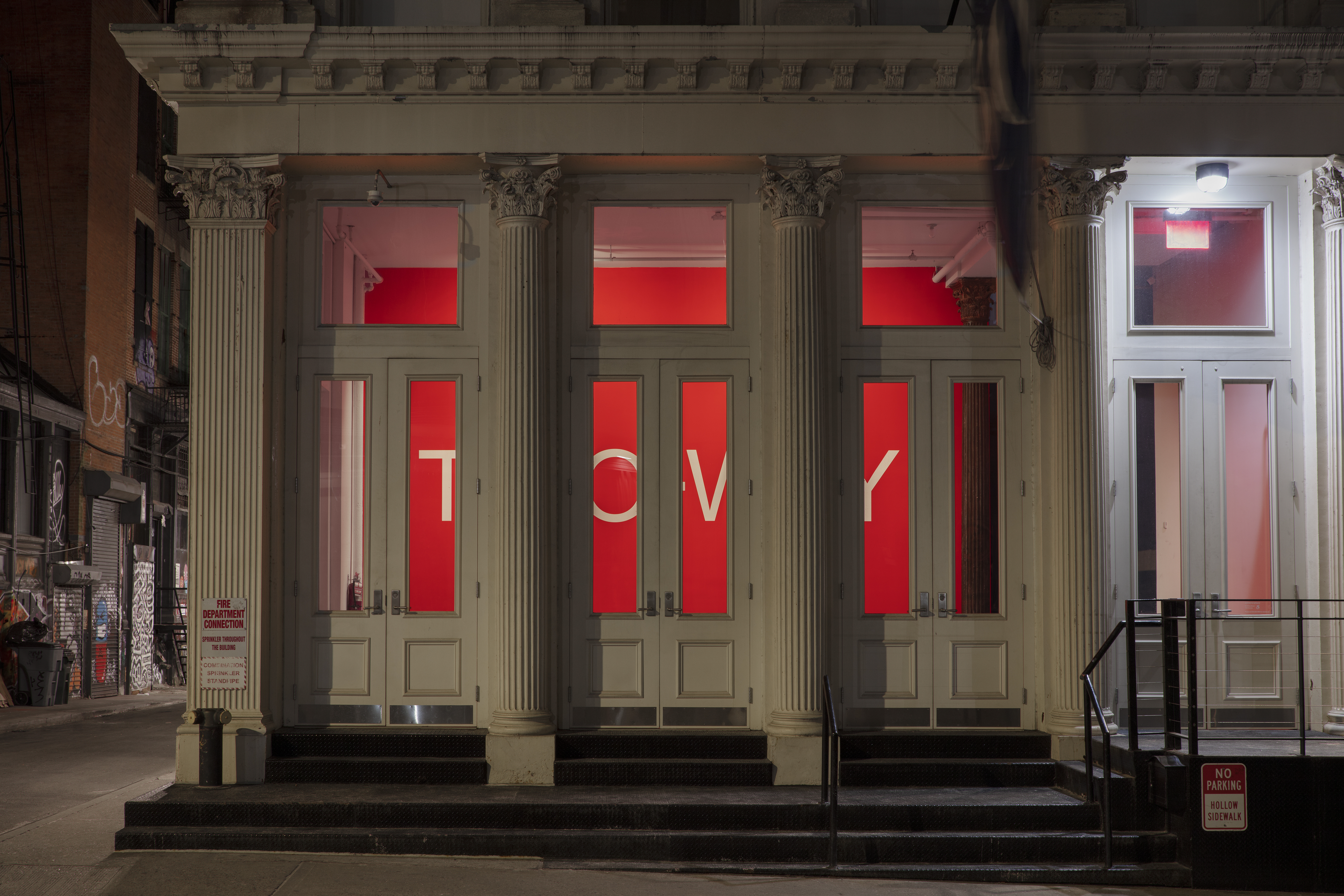 An exterior view of a gallery with white neoclassical columns and a row of three glass doors. Inside the gallery is a bright red wall with large white text: the text is partially obscured by the building’s columns and doorways.
