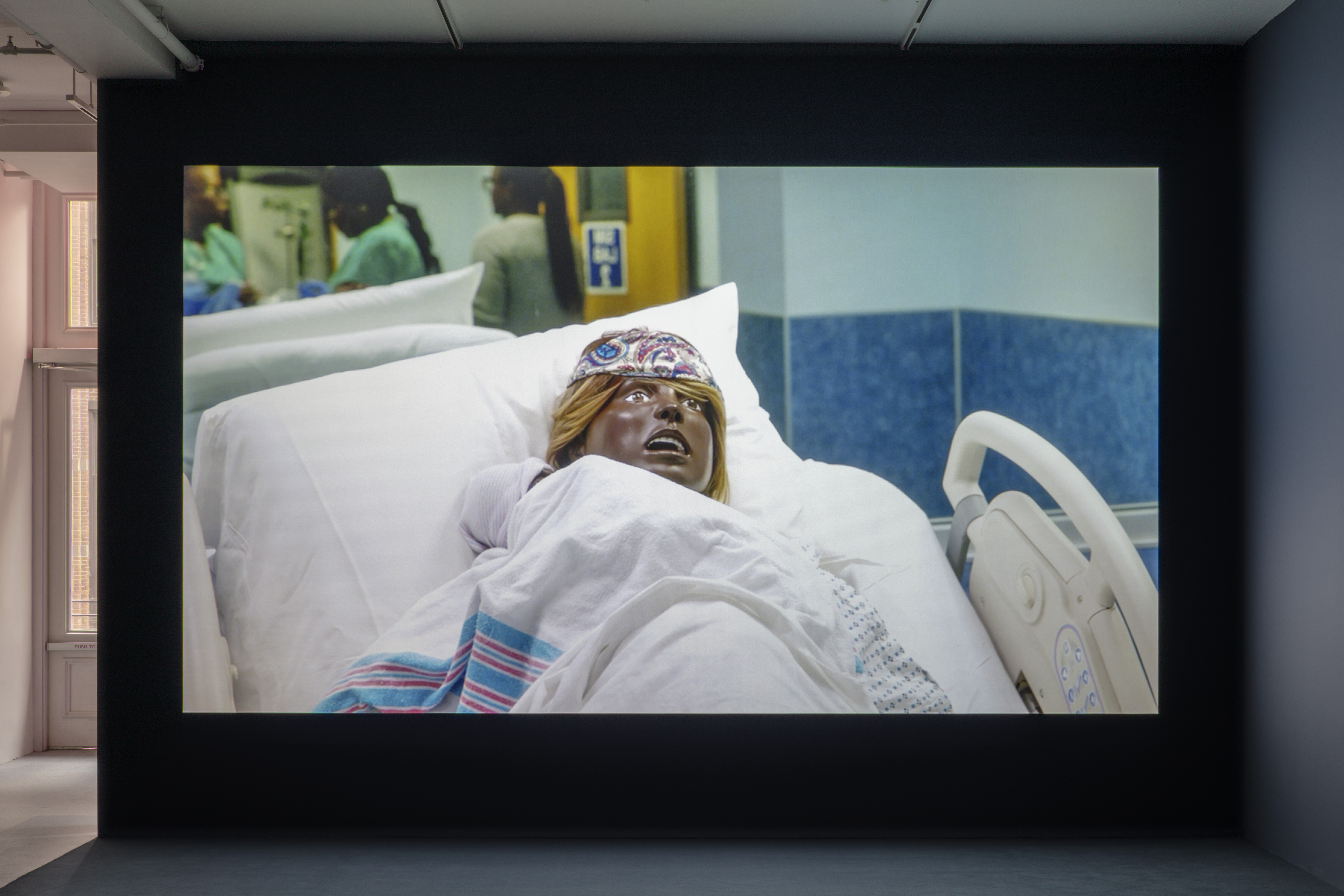 A darkened gallery space with a video projection centered on a dark blue wall. The projection depicts a close-up of a black birthing mannequin reclined on a hospital bed in a blue patient room. A newborn mannequin swaddled in a hospital receiving blanket lays on the birthing mannequin’s chest.