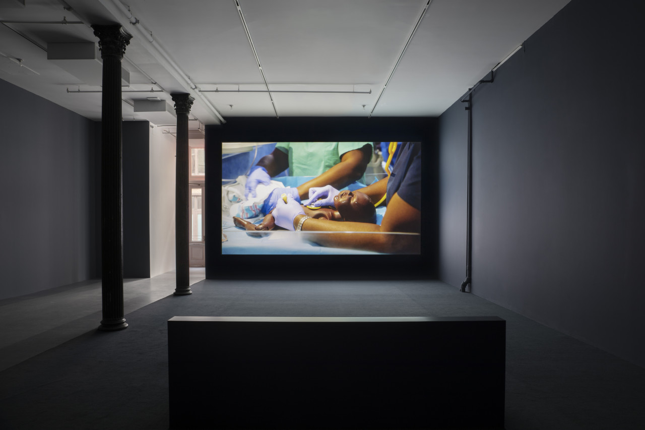 A darkened gallery space with dark blue walls, dark blue carpeting, and light blue linoleum tiling. In the foreground a bench is centered in front of a large wall projection. The projection depicts a close-up of a newborn black mannequin laying down, being tended to by medical professionals.