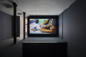 A darkened gallery space with dark blue walls, dark blue carpeting, and light blue linoleum tiling. In the foreground a bench is centered in front of a large wall projection. The projection depicts a close-up of a newborn black mannequin laying down, being tended to by medical professionals.