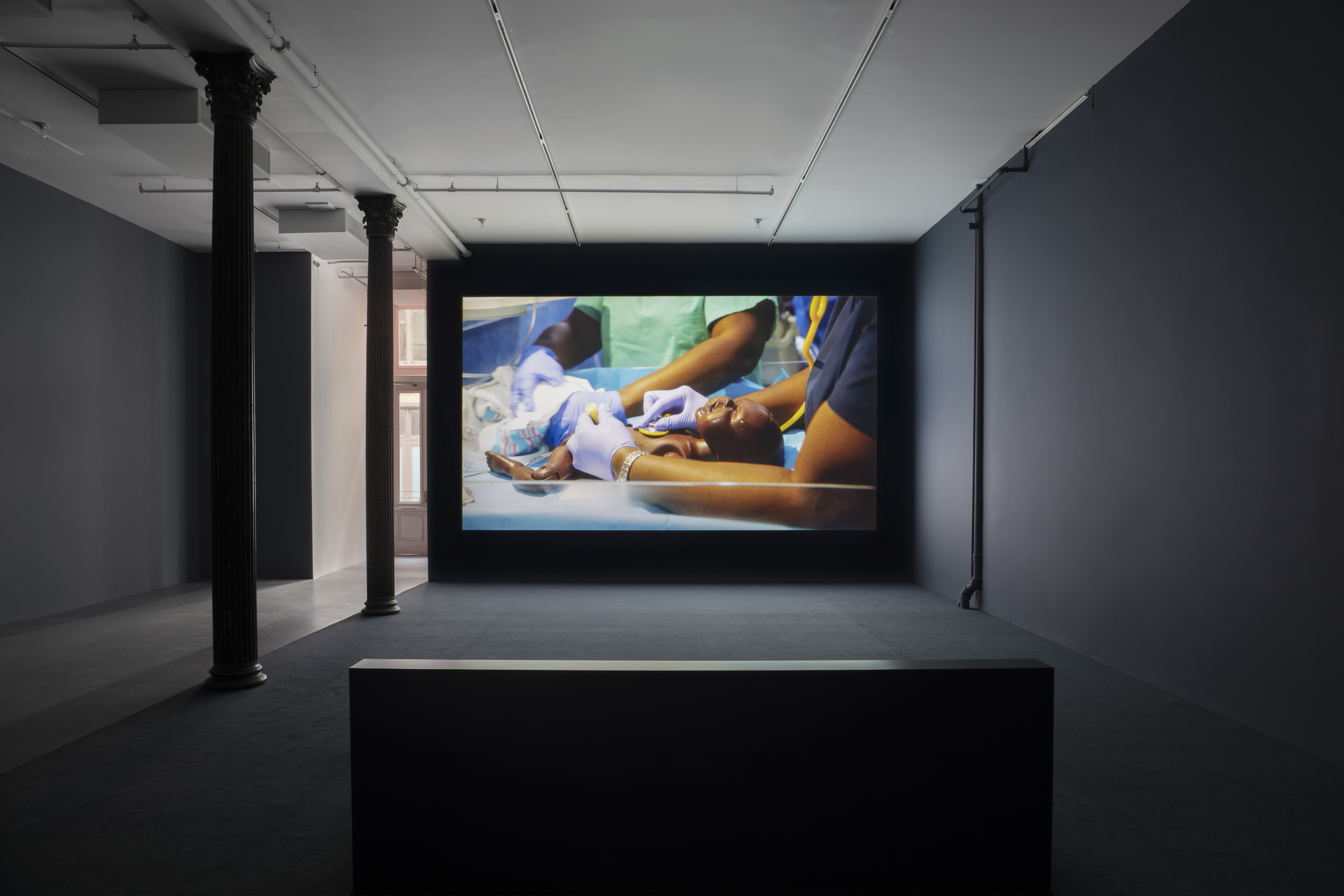 A darkened gallery space with dark blue walls, dark blue carpeting, and light blue linoleum tiling. In the foreground a bench is centered in front of a large wall projection. The projection depicts a close-up of a newborn black mannequin laying down, being tended to by medical professionals.