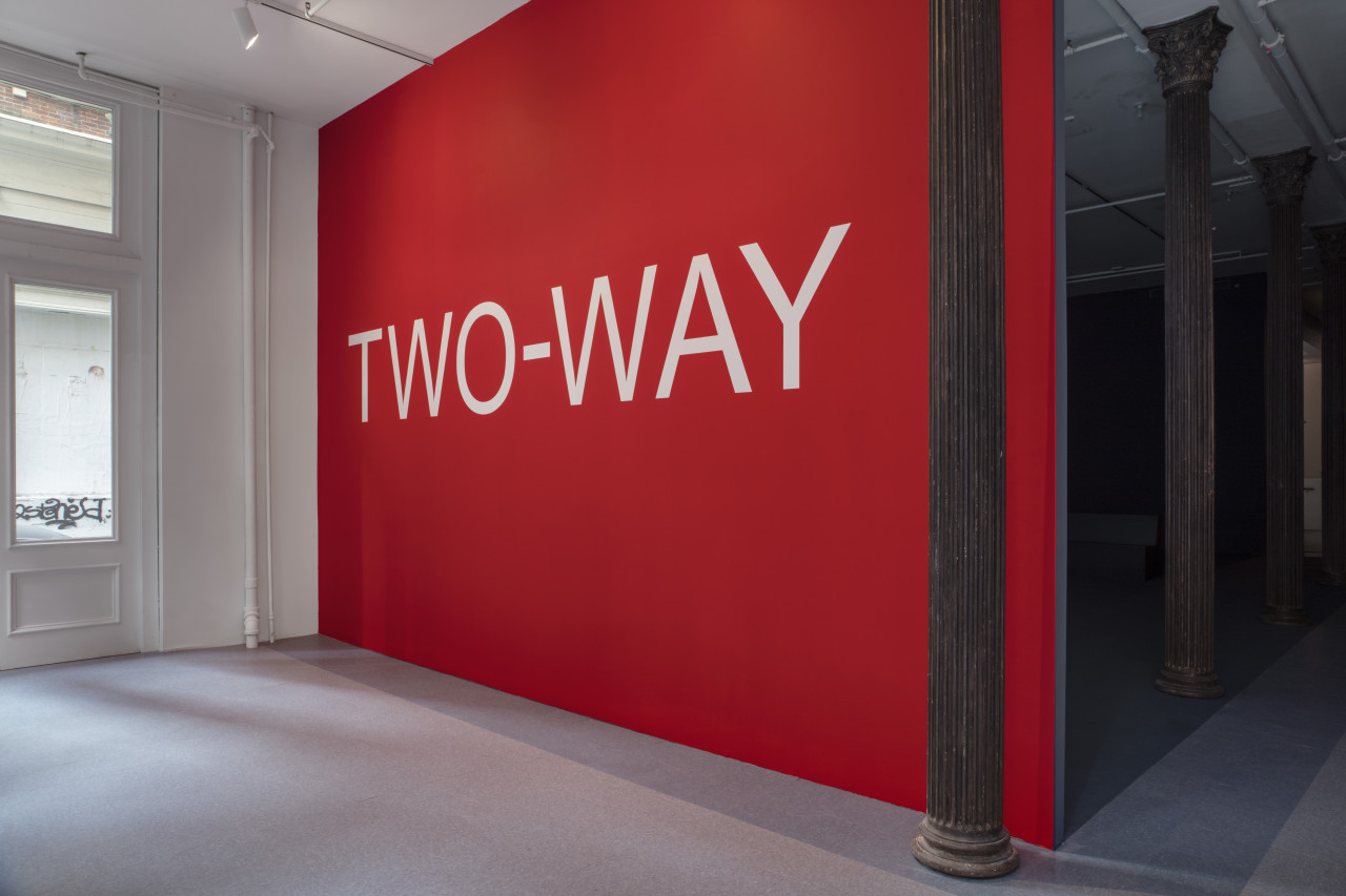 A brightly painted red wall with the phrase “TWO-WAY” centered in large white text. The floor in front of the wall, and to the side is covered in a pattern of light blue linoleum tiles. Brown columns stretch into the darkness behind the wall.