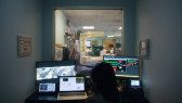 A small control room looks into an adjoining hospital patient room via an interior window. Inside the room a person sits in front of multiple display screens and observes the activity in the patient room.