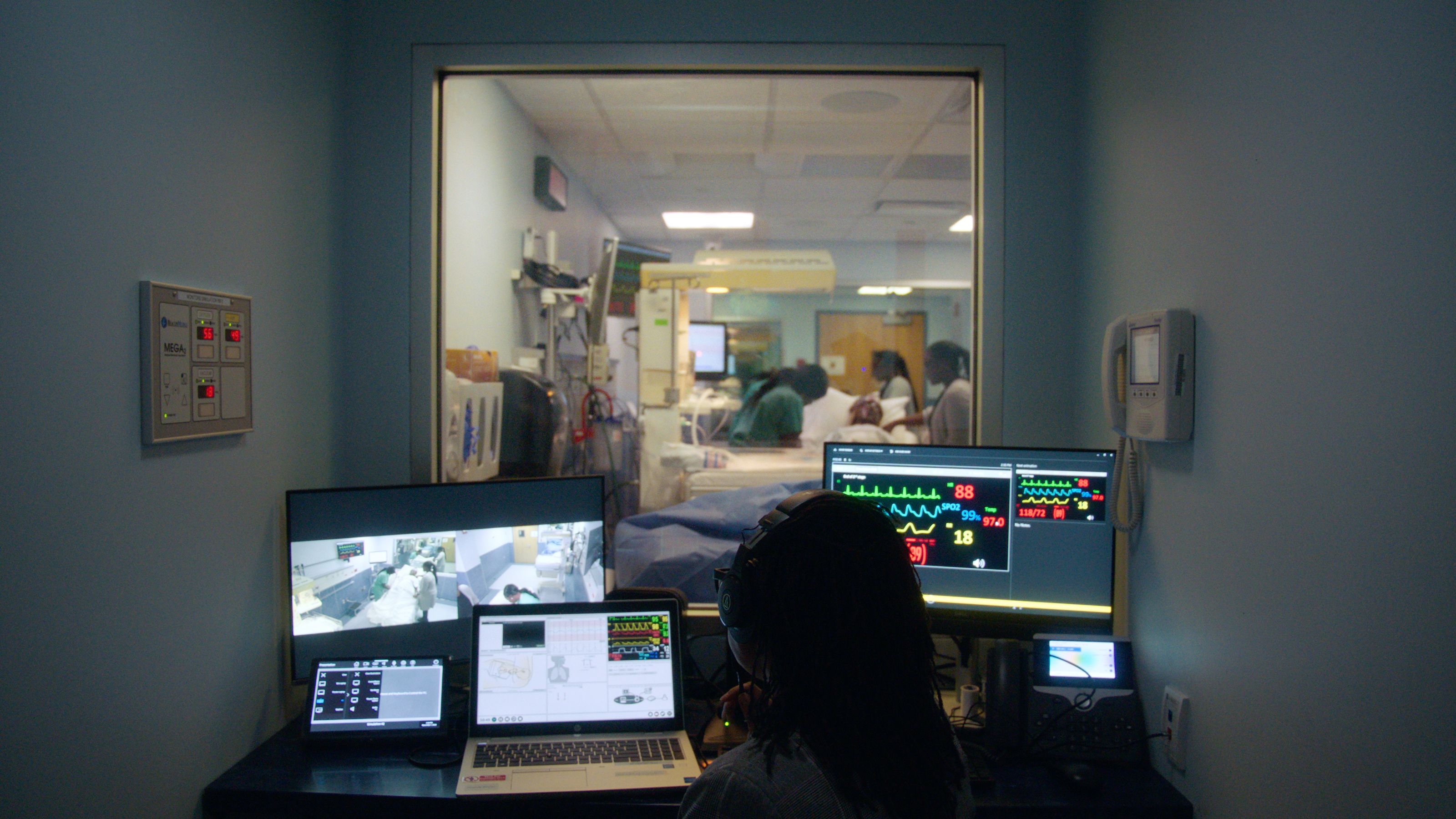A small control room looks into an adjoining hospital patient room via an interior window. Inside the room a person sits in front of multiple display screens and observes the activity in the patient room.