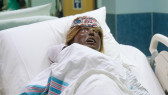 Close-up of an upright black birthing mannequin reclined on a hospital bed in a blue patient room. A newborn mannequin swaddled in a hospital receiving blanket lays on the birthing mannequin’s chest.