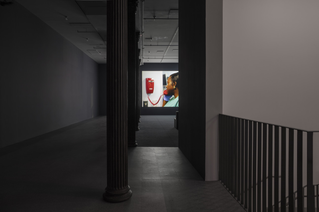 A darkened hallway leads into an open gallery space with dark blue walls, dark blue carpeting, and light blue linoleum tiling. In the background a video is projected onto the farthermost wall. The projection depicts a nurse in green scrubs using a red telephone affixed to the wall.