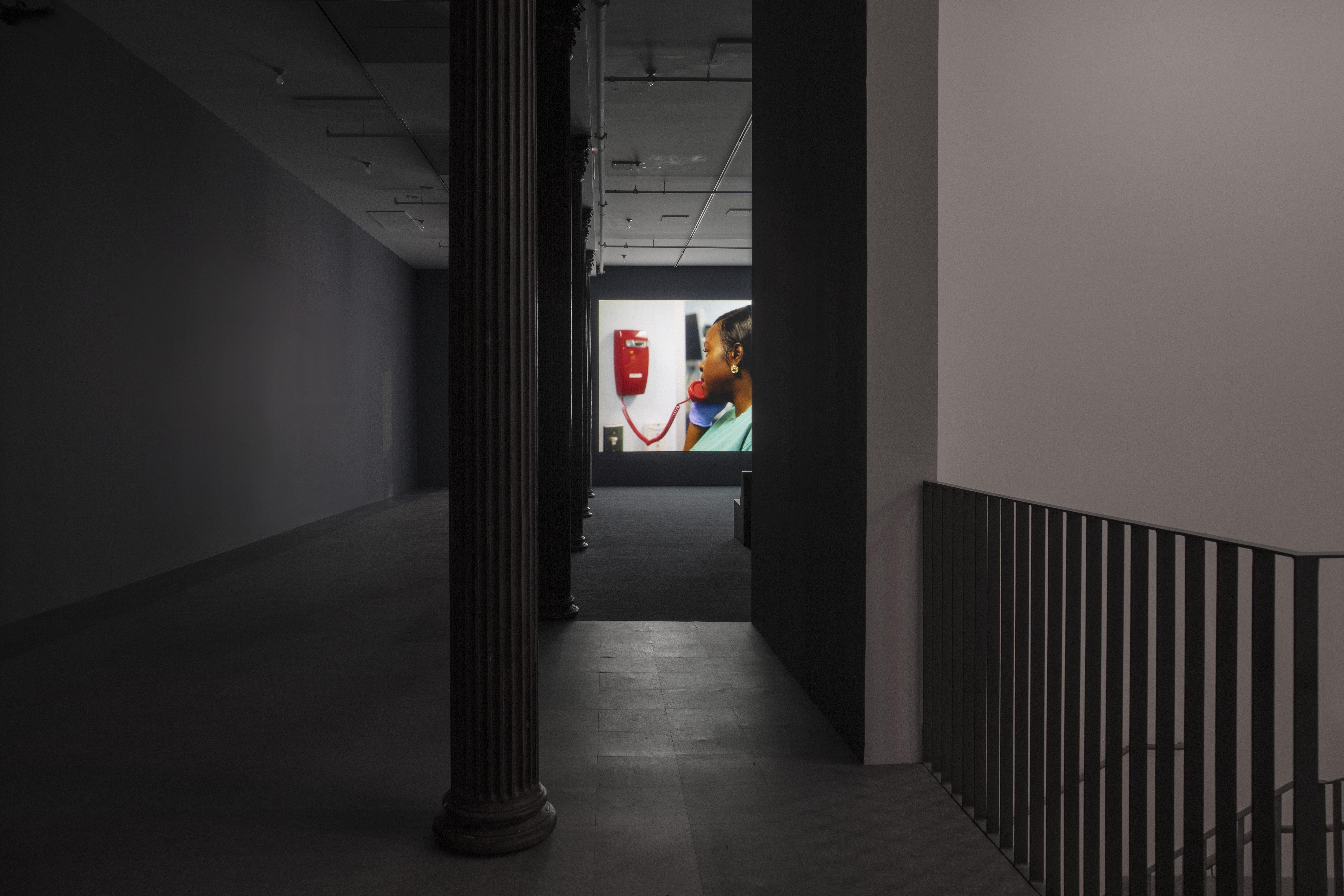 A darkened hallway leads into an open gallery space with dark blue walls, dark blue carpeting, and light blue linoleum tiling. In the background a video is projected onto the farthermost wall. The projection depicts a nurse in green scrubs using a red telephone affixed to the wall.