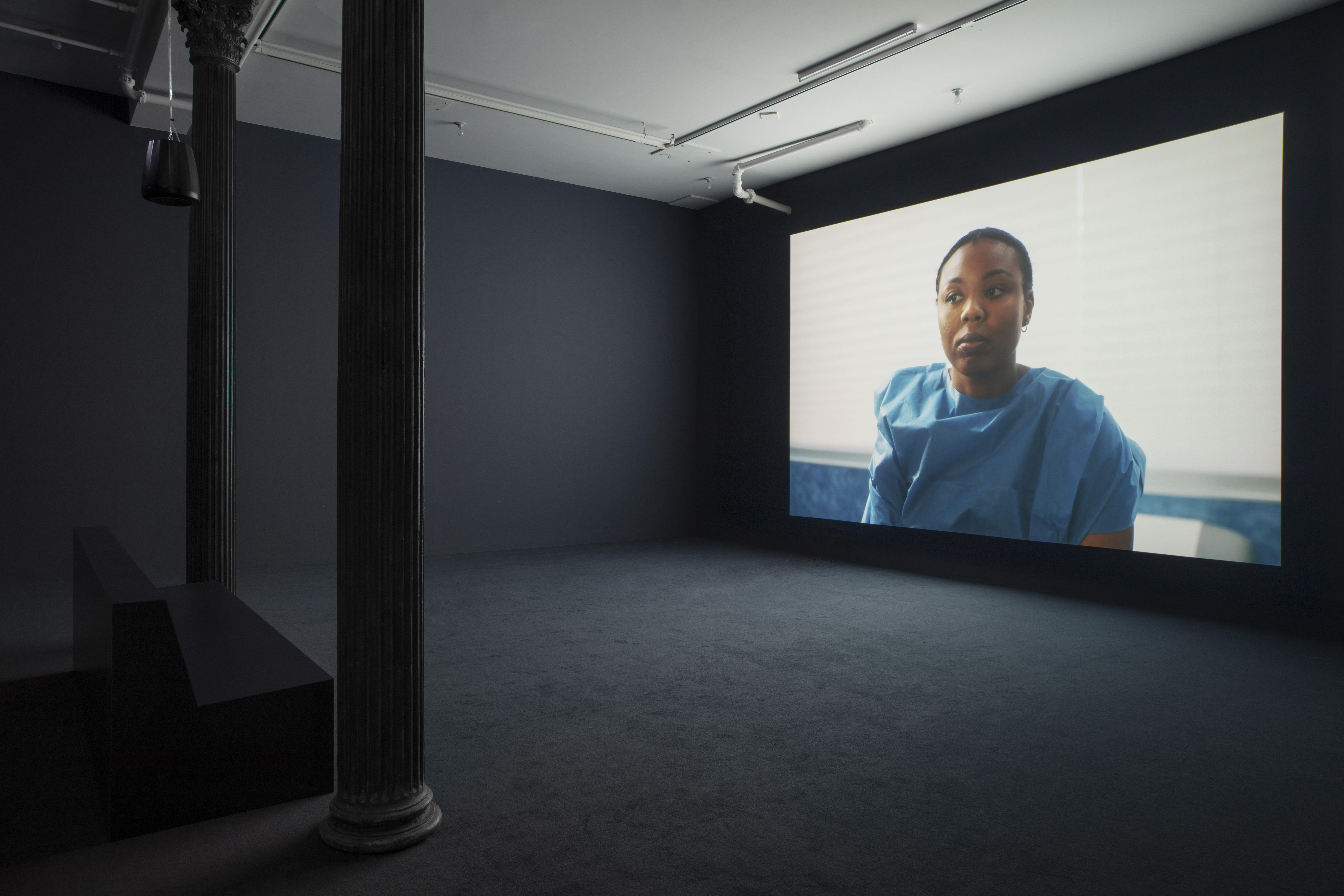 A darkened gallery space with dark blue walls and dark blue carpeting. On the left sits a bench, which faces a large wall projection on the right. The projection depicts a black patient in a blue hospital gown sitting on an examination table.