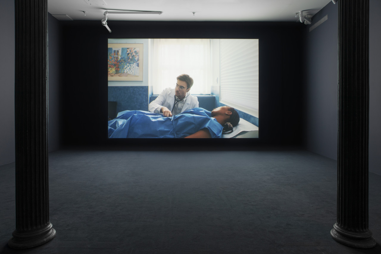 A darkened gallery space with dark blue walls and dark blue carpeting with a large wall projection. The projection depicts a black patient in a blue hospital gown laying down on an examination table. A white doctor holds a stethoscope to the patient’s stomach.