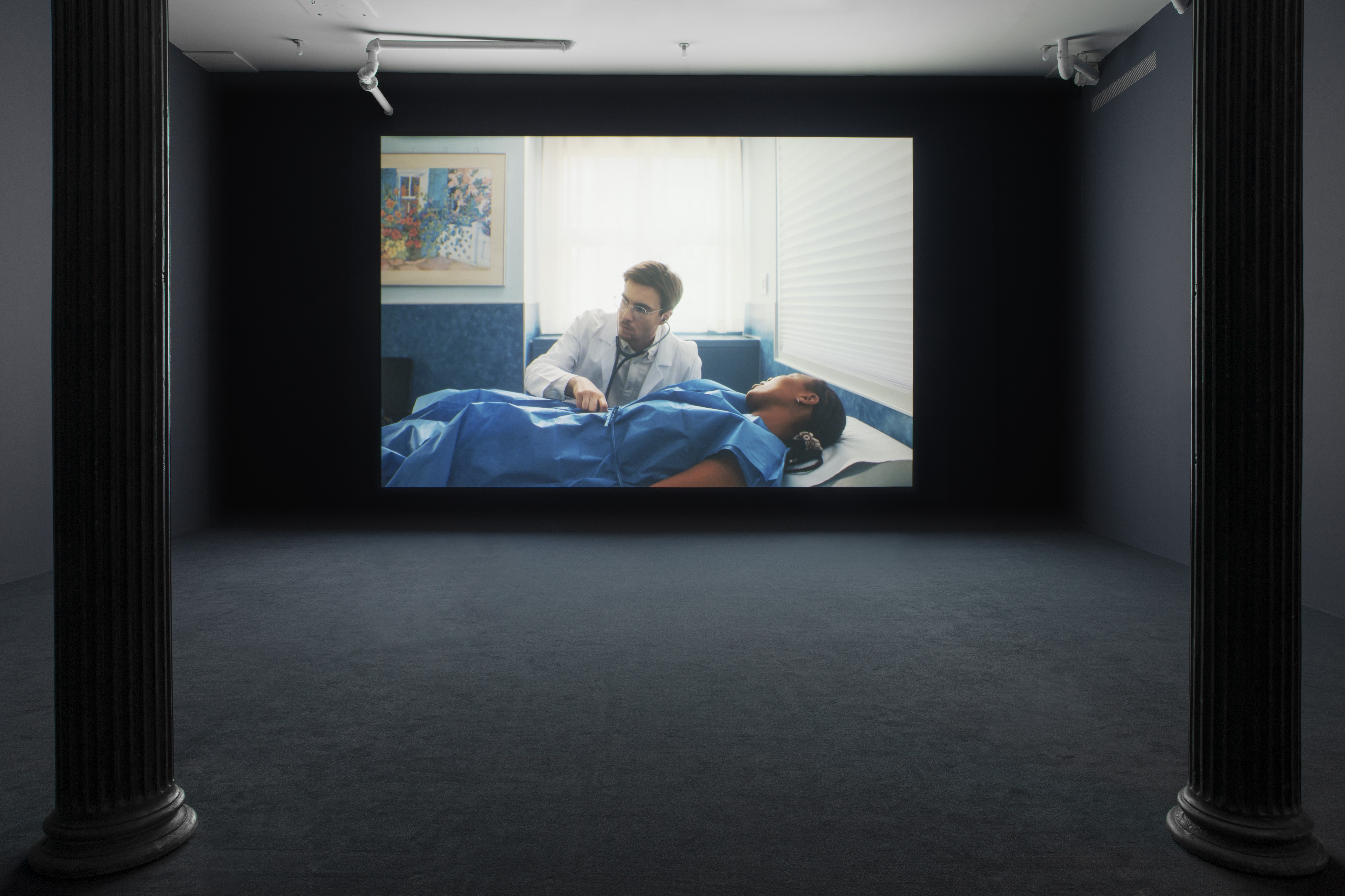 A darkened gallery space with dark blue walls and dark blue carpeting with a large wall projection. The projection depicts a black patient in a blue hospital gown laying down on an examination table. A white doctor holds a stethoscope to the patient’s stomach.