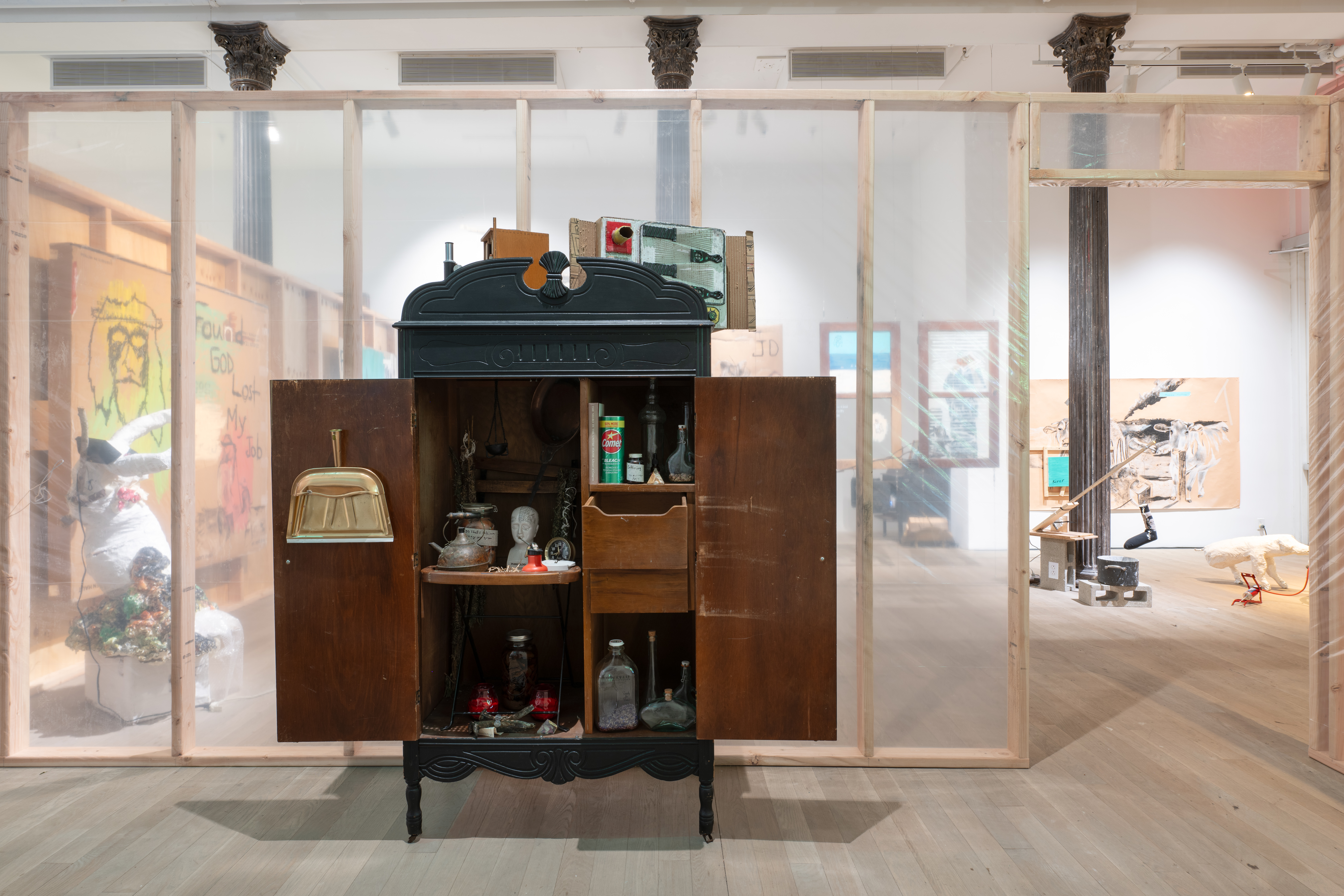 Color photo of an installation work. An antique cabinet with its doors open reveals shelves and drawers filled with household items such as jars, books, herbs, candles, a teapot, a cooking pan, cleaning products and a clock. On the top of the cabinet
