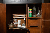 Color image of an old wooden cabinet, with the doors open revealing various objects placed on the shelves including: a black kettle, a glass jar with eggshells, a white bust, a clock, a fire alarm, a book, Comet bleach, and empty glass bottles