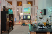 Color photo of a installation and sound work. In the right foreground, two lab tables contain a lab equipment such as beakers, distilling chambers, jars, hot plate and two blenders. On the left midground, an antique cabinet with its doors open, reveals shelves and drawers filled with more lab equipment, such as a microscope and household items such as a dustpan. In the background, two box television screens sit on top of wooden lab tables. A posterboard with a periodic table rests on top of the televisions. Each 