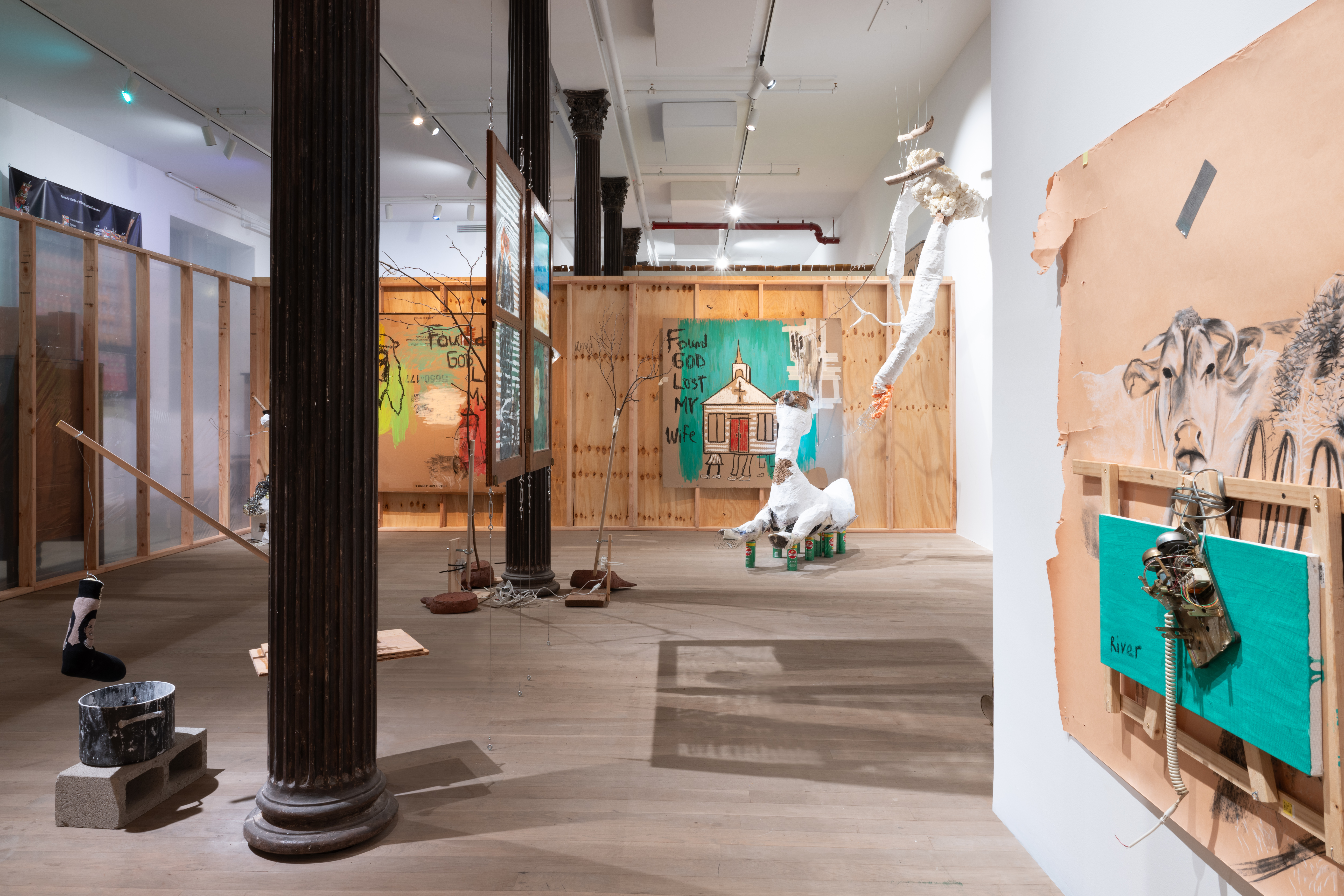 Color photo of an installation work. A row of structural columns cut through the installation. In the foreground, a kinetic scutlpure featuring a sock being dipped into a pot sit on the floor next to a column. Behind the first column, two windows with images painted on the glass hang from the ceiling. Behind the windows, ceramic sculptures and kinetic sculptures featuring branches surround another column. To the right of the ceramic sculptures and branch sculptures, large paper-mache sculptures rest on the floor and hang on the walls. In the right forground, a large drawing done on craft paper featuring a cow hangs on the wall. In the background, two large drawings done on cardboard are hung from a wooden partition wall.
