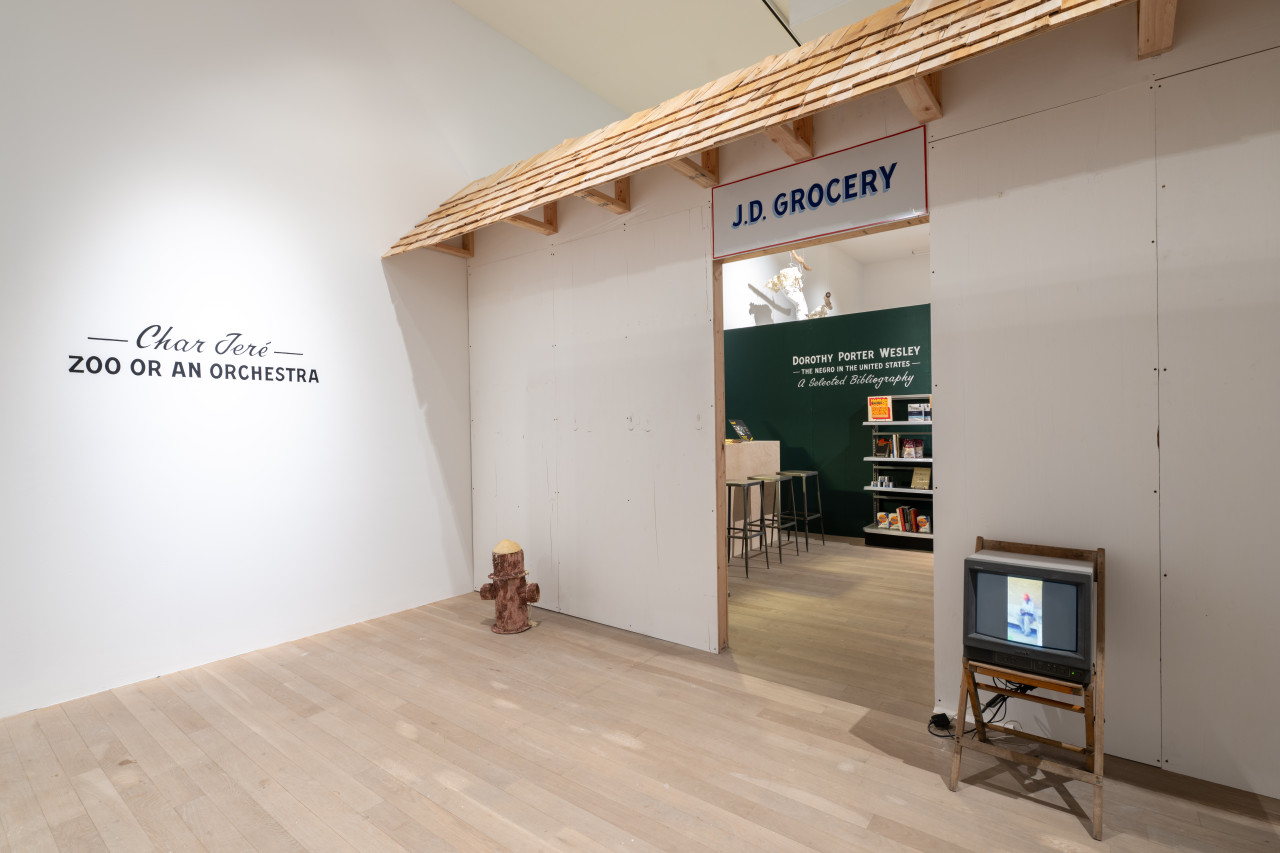 Color photo of the enterance into the exhibition. A sign reading: "J.D. Grocery" is placed above a doorway. A wooden awning meant to look like a roof of a building goes across the width of the partition wall. The doorway is flanked by a cardboard fire hydrant and a box television on a small wooden chair. Through the doorway, three bar stools and a bookshelf with books and home goods are visable.
