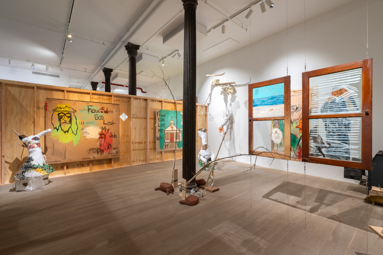 Color photo of an installation work in a gallery. On a wooden partition wall in the back of the space, two large carboard drawings hang. To the left, in front of the wooden parition, a paper-mache sculpture is placed on the floor. In the center of the image, ceramic works and kinetic sculptures made of branches surround a structural column. Behind the column, there are more paper-mache sculptures placed on the floor and hanging off the wall. In the right foreground, two windows with images painted on the glass hang from the ceiling.