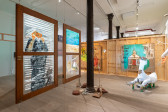 Color photo of an installation work in a gallery space. In the left foreground, two windows with images painted on the glass are hung from the ceiling. In the center midground, ceramic sculptures and kinetic sculptures made of branches surround a structural column. To the right of the column, a large paper-mache sculpture rests atop Comet cleaner bottles. In the background, two large drawings done on carbaord hang from a wooden partition wall.