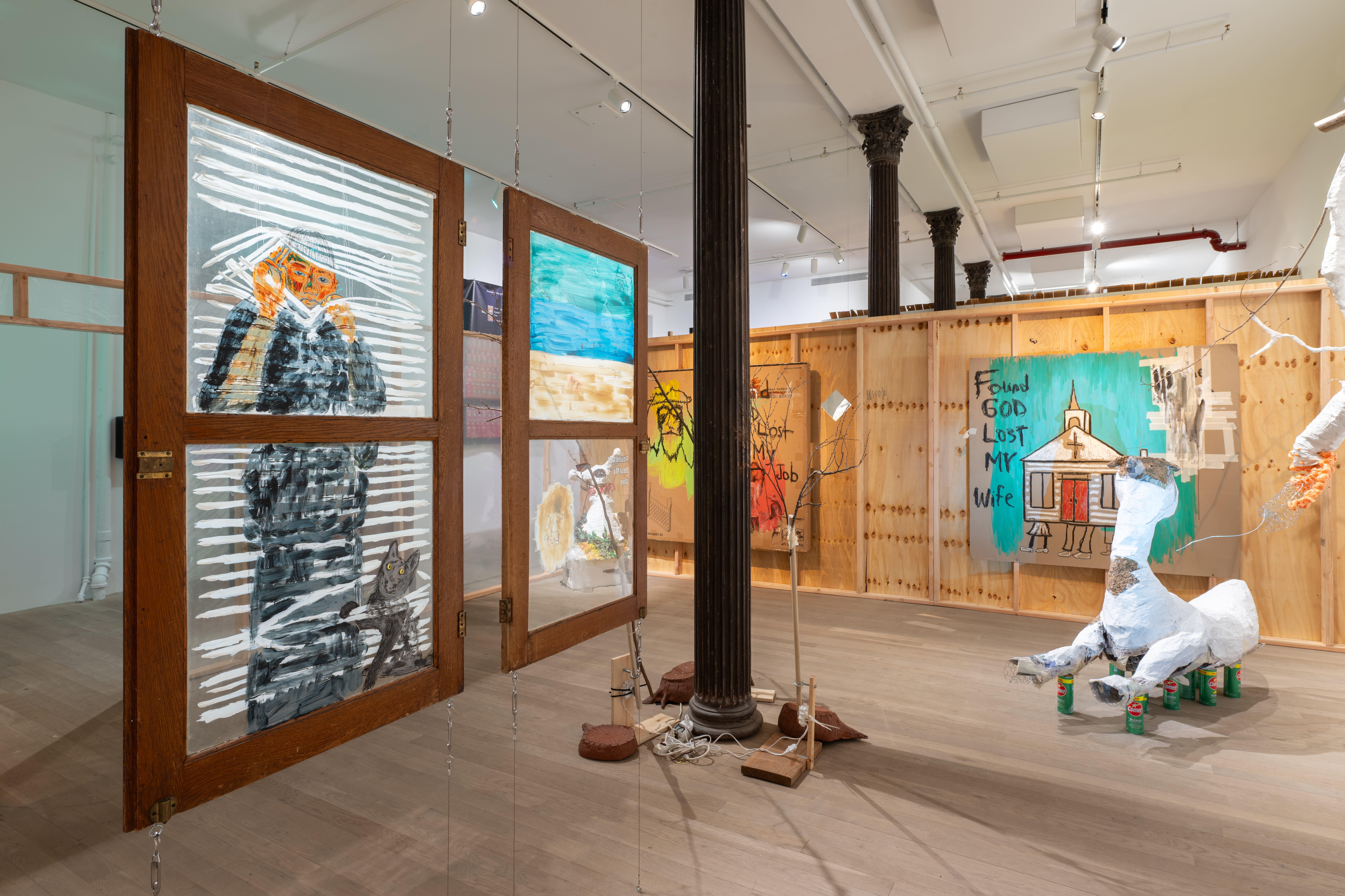 Color photo of an installation work in a gallery space. In the left foreground, two windows with images painted on the glass are hung from the ceiling. In the center midground, ceramic sculptures and kinetic sculptures made of branches surround a structural column. To the right of the column, a large paper-mache sculpture rests atop Comet cleaner bottles. In the background, two large drawings done on carbaord hang from a wooden partition wall.