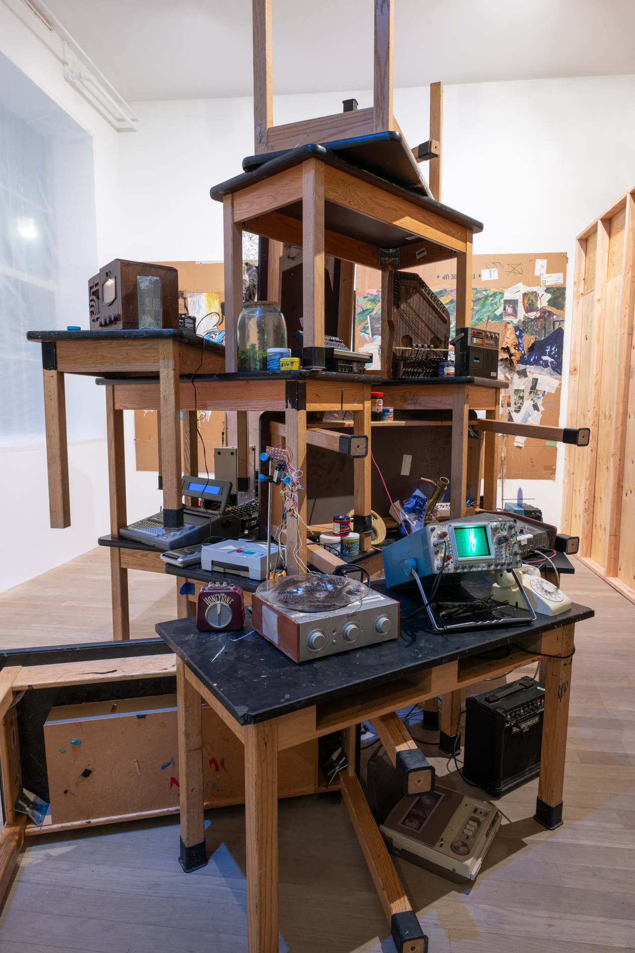 A color photo of an installation and sound work in a gallery space. Wooden tables with black table tops are stacked upon eachother, creating a structure of tables about 20 feet high. The tables are densely packed and populated by a variety of insturments, automated sound objects and other found items such as hair care products and medicine bottles. Behind the stack of tables, a large piece of cardboard hangs on the wall, filled with drawings and collaged elements.