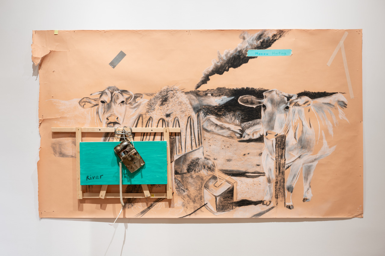 Black and white drawing of two cows. A tv sits on the floor between them, and a smoke column is seen in the background. A blue piece of tape with the words Moooo hurting is taped on top of the smoke column. A wooden structure, an electronic device, and a small canvas painted in blue with the word River written across it, cover part of the cow drawing on the left.