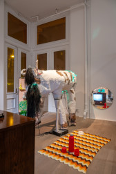Color photo of an installation work. A large, paper-mache sculpture of a cow-like creature stands next to a crocheted blanket laid on the floor. On top of the blanket there is a red thermos with two red mugs and a miniature white couch. On the back wall behind the paper-mache sculpture and blanket is a round mirror. The mirror contains a reflection of a large box TV with a static screen. On the left-side background, behind the paper-mache sculpture, two blow-dryers hang from a freestanding metal fixture.