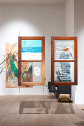 Color photo of an installation work in a gallery. Two windows with images painted on the glass hang from the ceiling. A branch emerges from the left edge of the image. Behind the windows, a colorful drawing on craft paper hangs on the wall.
