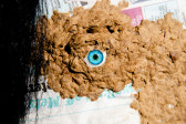 Color image of a piece of matted brown material, placed on top of a newspaper. In the center of the matted material, a glass eye with a bright blue iris is left uncovered. The left edge of the image is obscured by strands of black hair hanging down.