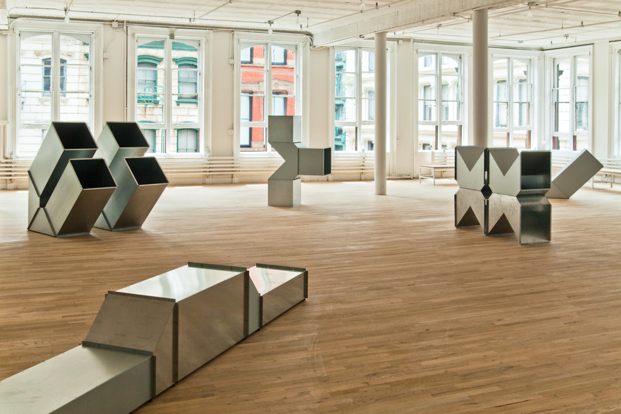 Four geometric, silver metal sculptures are installed on the floors of a light-filled gallery space. The sculptures are modular, hollow, and consist of cube-like and triangular metal components.