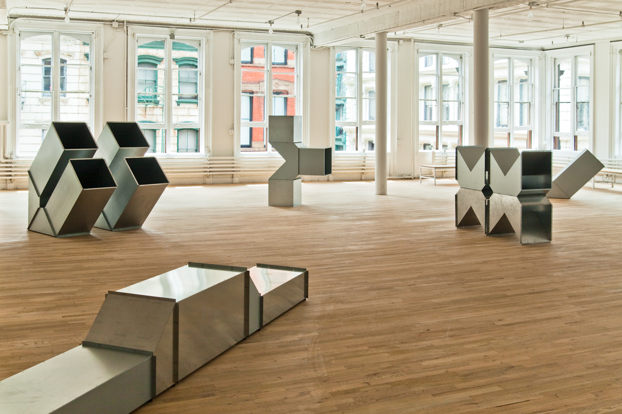 Four geometric, silver metal sculptures are installed on the floors of a light-filled gallery space. The sculptures are modular, hollow, and consist of cube-like and triangular metal components.