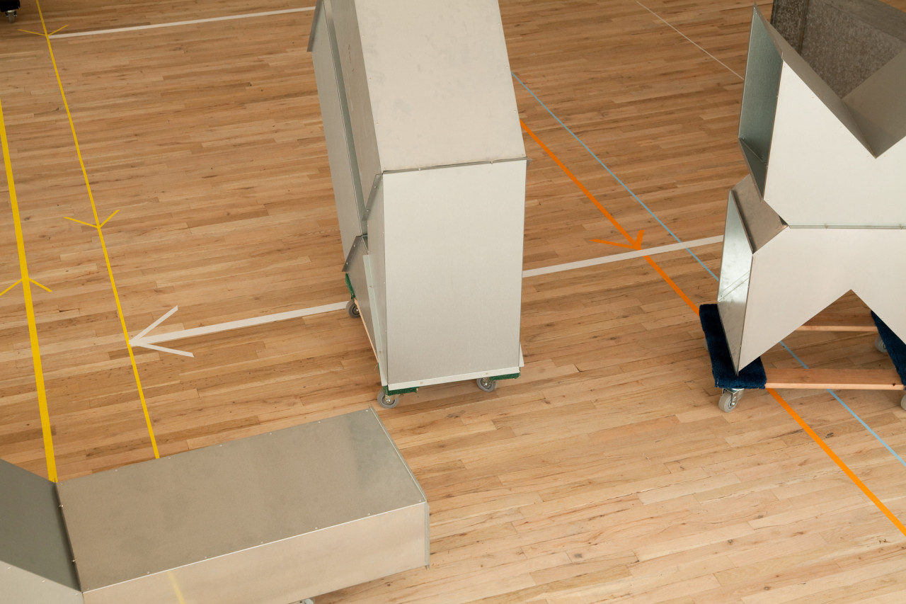 Three geometric, silver metal sculptures sit on low dolleys with wheels on a wooden gallery floor. The sculptures are modular, hollow, and consist of cube-like and triangular metal components. Long arrows of colorful tape are installed on the gallery floors.