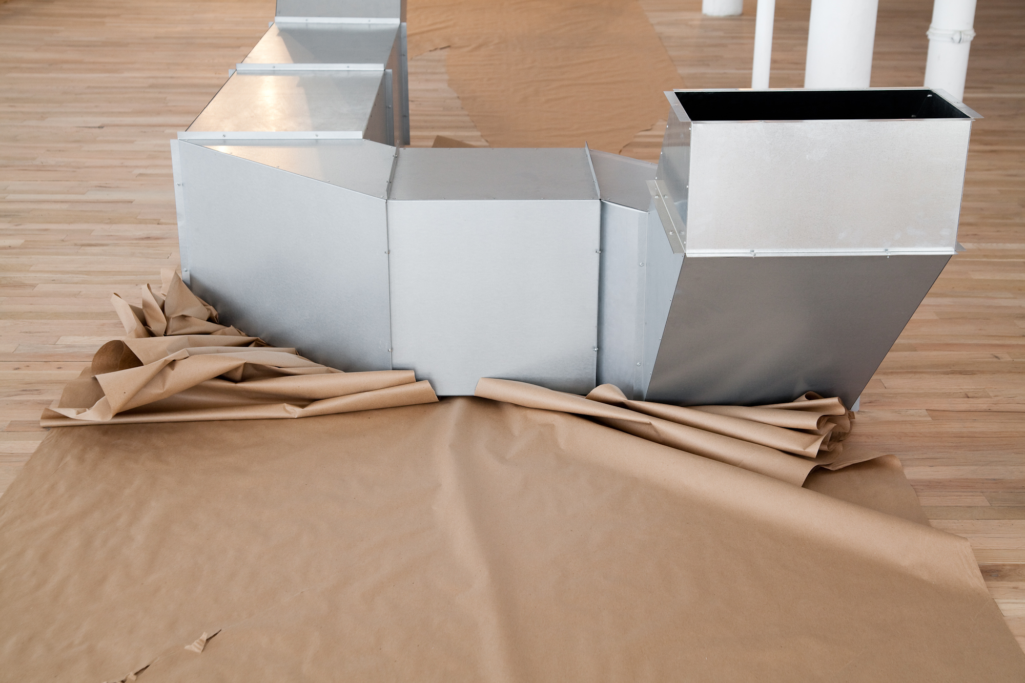 A geometric, silver metal sculpture sits on a wooden gallery floor. The sculpture is modular, hollow, and consists of cube-like and triangular metal components arranged in a long form that snakes across the ground. A large sheet of brown paper rests on the floor before the sculpture, crumpling against the sculpture's lower edge.