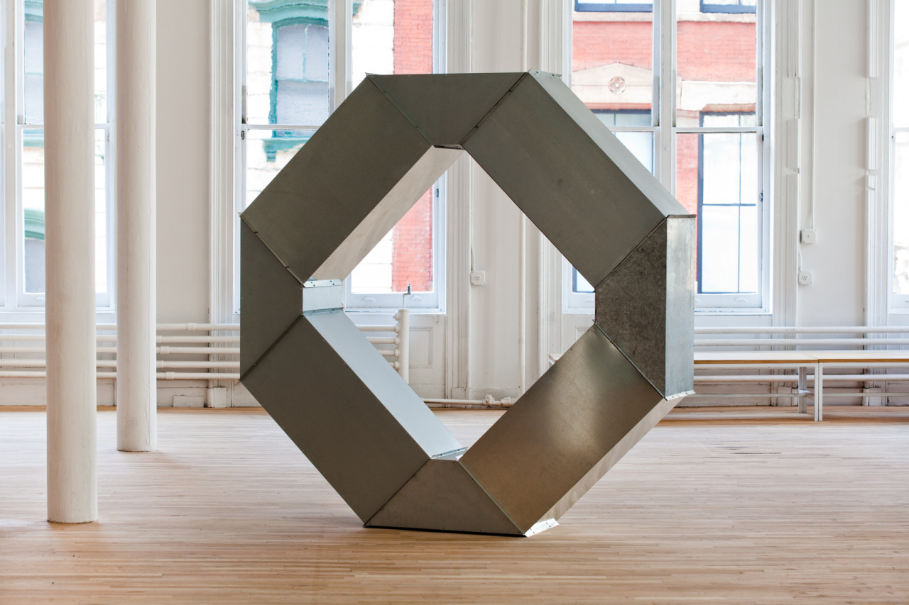 A geometric, silver metal sculpture sits on a wooden gallery floor in a brightly-lit gallery space. The sculpture is modular and consists of rectangular and triangular metal components arranged in a tall, ring-shaped structure.