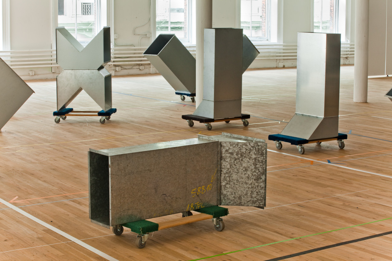 Six geometric, silver metal sculptures installed on low dolleys with wheels sit, scattered, across a wooden gallery floor. The sculptures are modular, hollow, and consist of cube-like and triangular metal components. Long arrows of colorful tape are installed on the gallery floors.