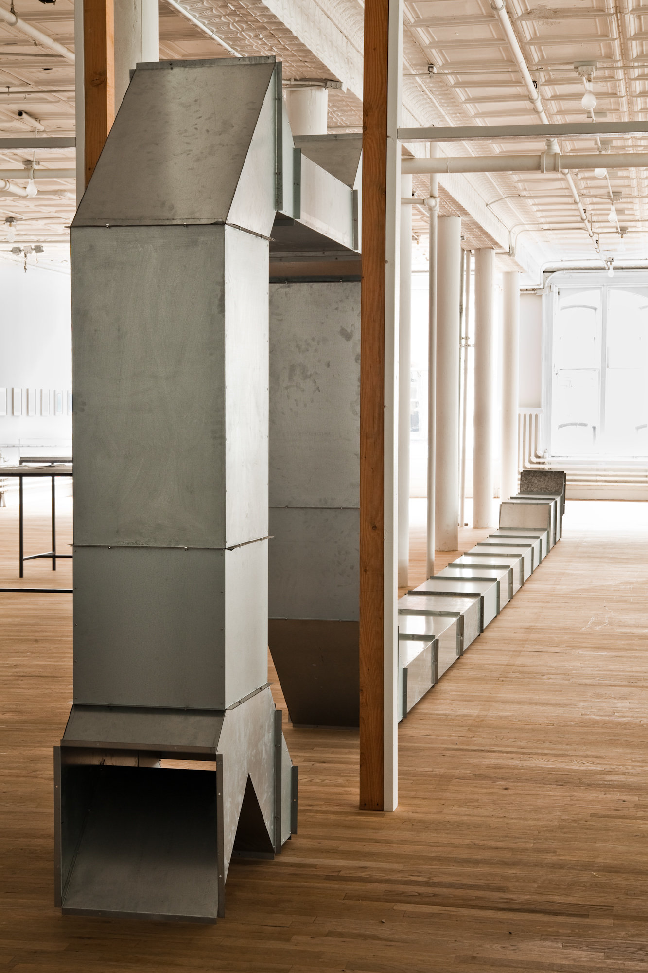 A geometric, silver metal sculpture sits on a wooden gallery floor. The sculpture is modular and hollow and consists of rectangular and triangular metal components arranged in a long form that snakes across the ground before arching up and down, forming a large U-shaped archway.
