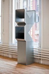 A geometric, silver metal sculpture sits on a wooden gallery floor in front of a large, light-filled window. The sculpture is modular, hollow, and consists of rectangular and triangular metal components. Two vent-like openings in the sculpture face the viewer, while the opposite side of the sculpture bends towards the gallery window.