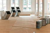 Several geometric, silver metal sculptures sit on a wooden gallery floor in a light-filled exhibition space. The sculptures are modular, hollow, and consist of cube-like and triangular metal components. At the photo's center, a long roll of crinkled brown paper enfurls across the floor of the gallery, resting in between the sculptures.