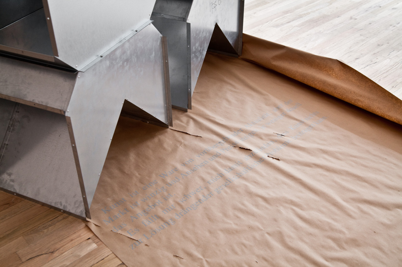 A geometric, silver metal sculpture sits on a roll of brown paper spread across a wooden gallery floor. The sculpture is modular and consists of triangular metal components. Gray text, in German, is subtly printed across the paper. Two long rips appear in the paper