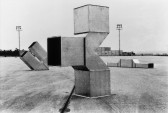 Black and white photograph of three, geometric metal structures sitting on a wide, outdoor concrete plaza. One of the metal structures is situated in the center of the photograph, while the additional two sit behind it, flanking it on either side. The structures are composed of hollow cube-like and triangular components.