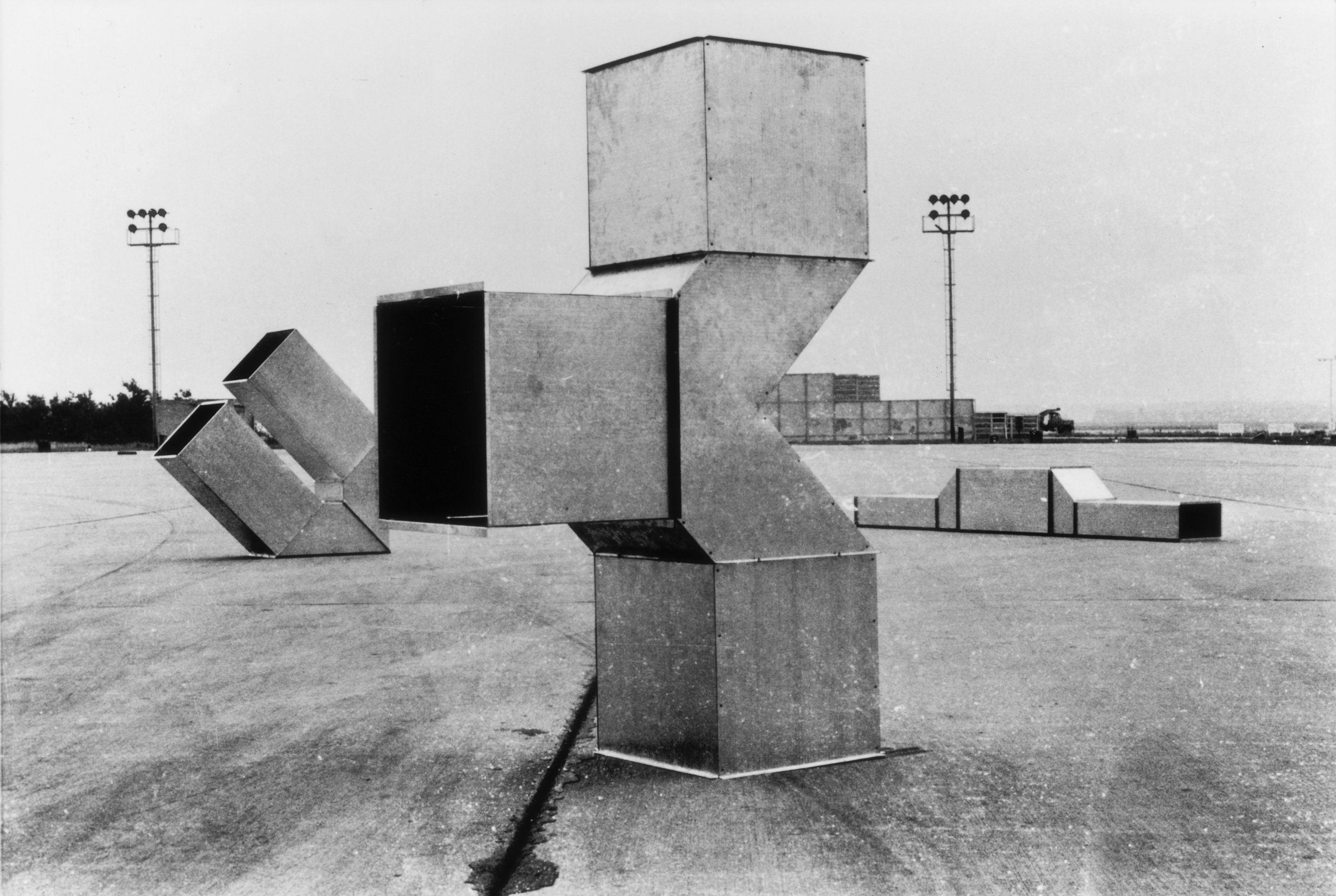 Black and white photograph of three, geometric metal structures sitting on a wide, outdoor concrete plaza. One of the metal structures is situated in the center of the photograph, while the additional two sit behind it, flanking it on either side. The structures are composed of hollow cube-like and triangular components.