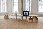 A long rectangular wooden table with three TV monitors facing outwards on different sides. Six small wooden seats with wired headphones resting on them are arranged in front of the monitors. One visible screen displays an image of a man sitting at a table.