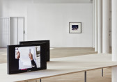 Two black monitors facing opposite directions sit on a large, rectangular wooden table. One screen is visible, showing an image of two hands holding a piece of white paper with typed text. In the background, a framed photograph is installed on a white gallery wall.
