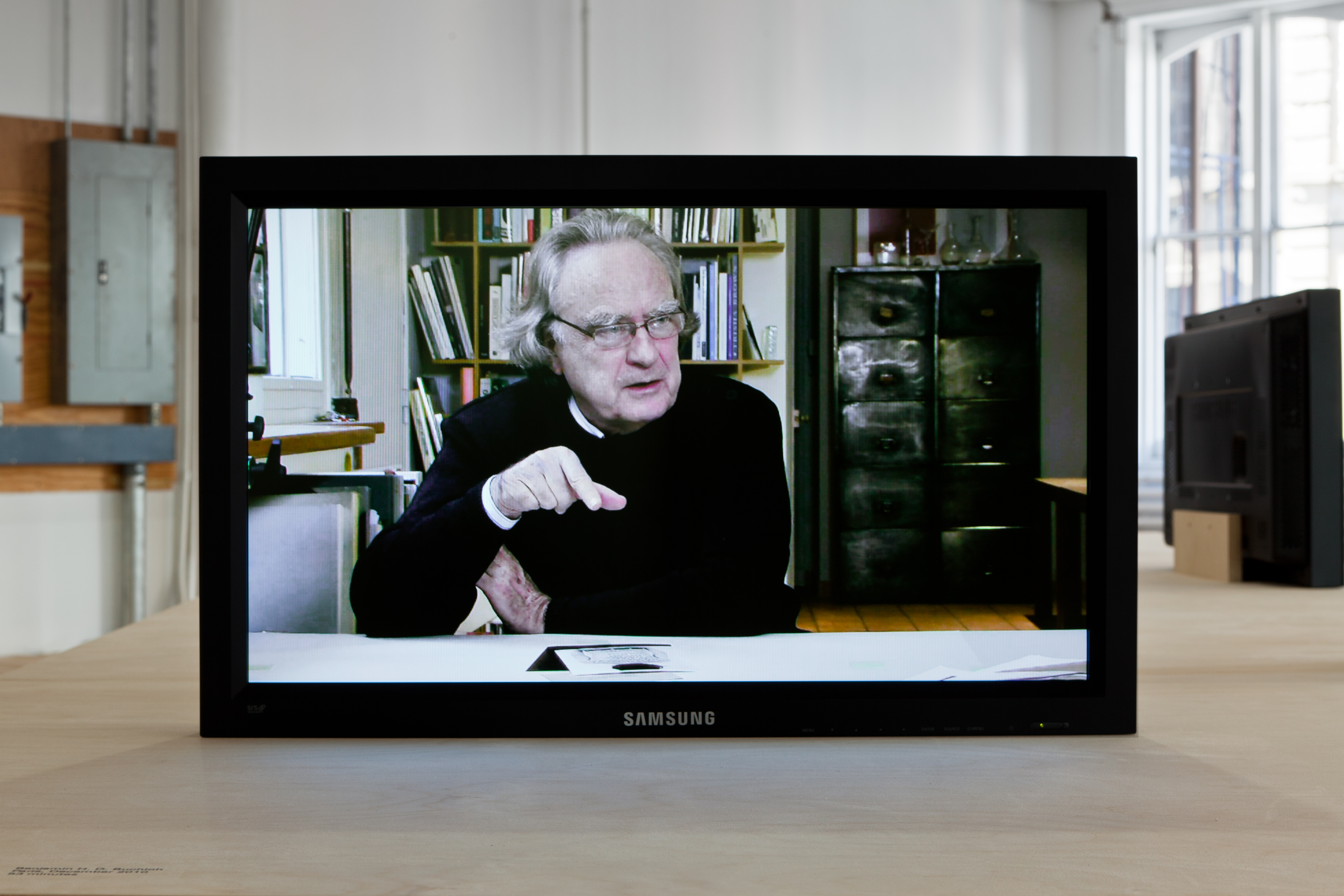 A Samsung television monitor displaying Benjamin Buchloh, in a black sweater and thin rectangular glasses, being interviewed.