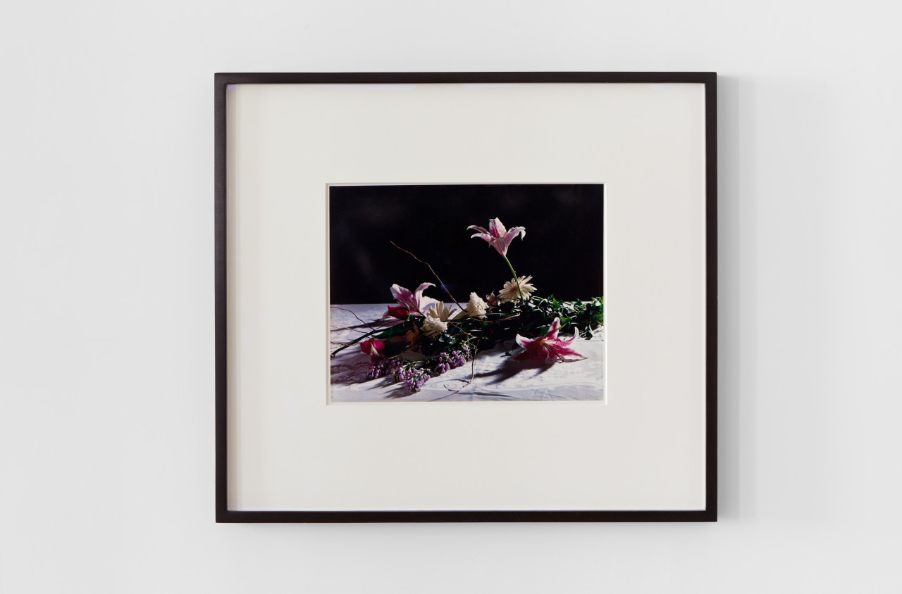 A framed photograph displayed on a white wall. The photograph shows multicolored flowers laying on a white surface with a black background.