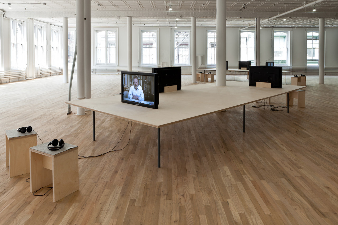 A long rectangular wooden table supporting three television monitors facing outwards on different sides. Two small wooden seats with wired headphones resting on them are arranged in front of the leftmost monitor.