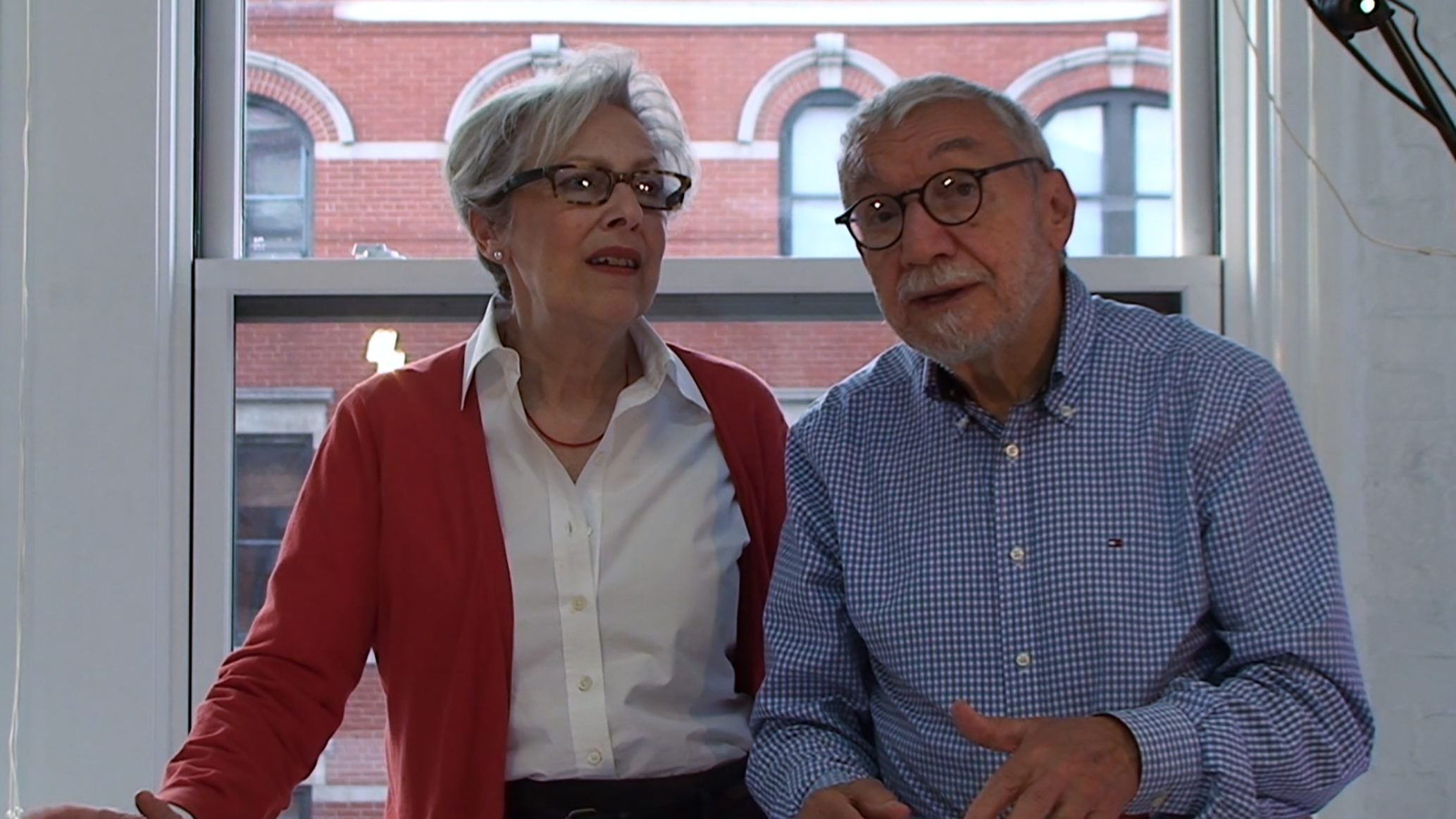 Two people in the middle of a conversation. The woman is wearing a red cardigan, a white collared shirt, and has short gray hair with square black glasses. The man is wearing a checked blue and white collared shirt and has gray facial hair and round black glasses.